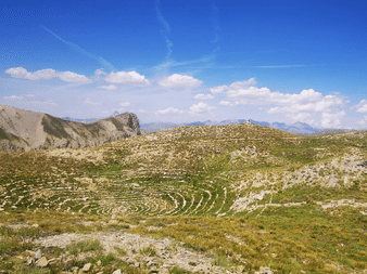 Højalpine eng med cirkulære stenmønstre i Queyras. Klippefyldte bjergtinder rejser sig under blå himmel med hvide skyer.