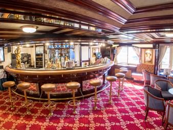 Luksuriøst bar-interiør med buet træ-disk, barstole, rødt mønstret tæppe og siddeområde med lænestole og vinduer.