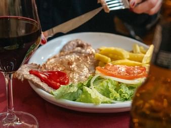 Plate with grilled meat, fries, lettuce, tomato and red pepper beside a glass of red wine on a red tablecloth. Portuguese Camino day 10.