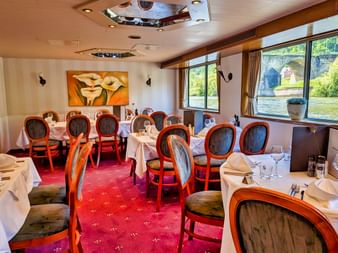 Elegant restaurantinteriør på MS Olympia med hvide duge, træstole med mørk polstring, rødt tæppe og store vinduer.