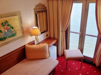 Skibskahyt med beige sofa, træskrivebord, spejl, bordlampe, indrammet kunst, gyldne gardiner og balkondør på MS Prinzessin Katharina.