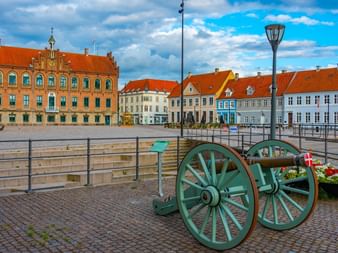 Nyborg slott med röd tegelfasad och färgglada stadshus kantar ett kullerstenstorg. En grön historisk kanon står i förgrunden.