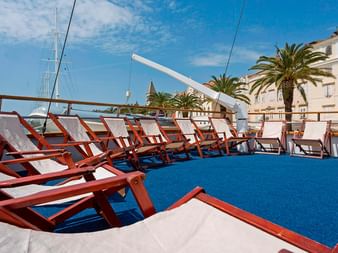 Træ liggestole med hvide hynder på blåt skibsdæk. Kroatisk kystby med palmer og historiske bygninger synlige.