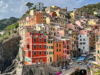 Colorful houses stacked on hillside in Cinque Terre, Liguria. Buildings in yellow, pink, orange and red overlook a small harbor with boats.