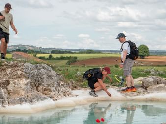 Three hikers with backpacks at a shallow water pool surrounded by rocks. Rural Italian landscape with fields and hills in the background.