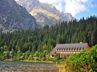 Stor bjerglodge med rødbrun facade ved en sø, omgivet af tæt nåleskov med klippefyldte Tatra-bjergtoppe i baggrunden.