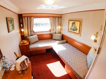 Kompakt skibskahyt med to enkeltsenge, vindue med gardiner, træmøbler og champagneservice på MS Prinzessin Katharina.