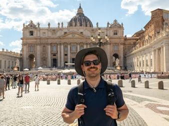 Man with backpack and hat standing in St. Peter's Square, Rome. St. Peter's Basilica with its iconic dome visible in the background.