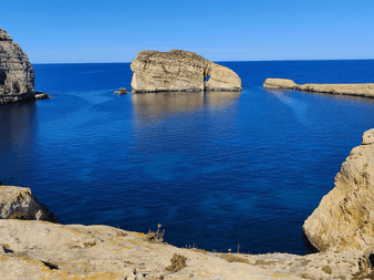 Dwejra Bay på Gozo, Malta, med dybt blåt hav omgivet af kalkstensklipper og markante klippeformationer under klar blå himmel.