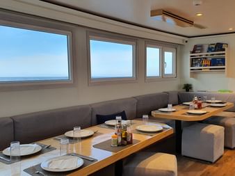 Indendørs spisestue på MV Tala med grå siddepladser, dækkede træborde og store vinduer med havudsigt.
