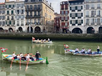 Three colorful traditional rowing boats with crews on green river water. Historic French buildings with shutters line the waterfront.
