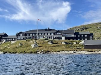 DNT Finsehytta hyttekompleks med mørke trebygninger og norsk flagg på steinete terreng ved et fjellvann under blå himmel.