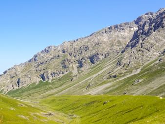 Bred grøn dal med vandrerute i Picos de Europa, Spanien. Stejle klippefyldte bjergskråninger rejser sig under klar blå himmel.