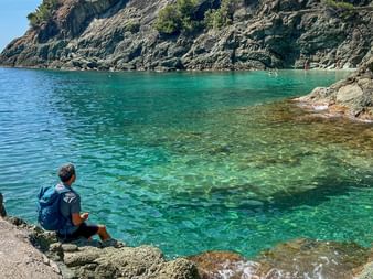 Hiker with backpack sitting on rocky shore overlooking turquoise bay in Cinque Terre, Liguria. Steep cliffs covered with vegetation frame the water.