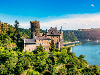 Middelalderligt slot med stentårne på skovklædt bakke over Rhinen nær Koblenz. Blå flod bugter sig gennem grøn dal under klar himmel.