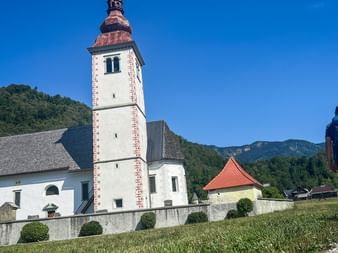 Vandrare med ryggsäck på grusväg bredvid vit kyrka med högt torn i Juliska alperna, Slovenien. Gröna berg under blå himmel.