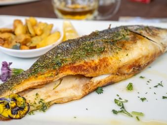 Grillet hel fisk pyntet med friske urter på hvid tallerken, serveret med ristede kartofler og spiselige blomster.