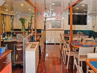 Interiør af MS Azimut salon med spiseborde med blå duge, hvide stole, en bar med flasker og grønne siddepladser ved vinduerne.