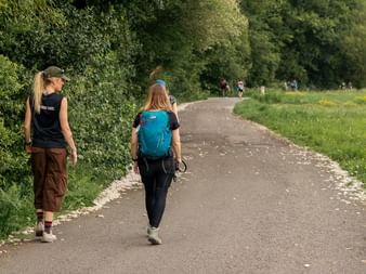 Two women walking on paved path through green countryside on Portuguese Camino. One wears blue backpack, trees and meadows line the trail.