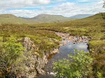 Stenet bæk løber gennem skotsk hedelandskab på West Highland Way med bølgende bakker og bjerge under skyet himmel.
