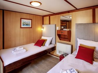 Dobbeltkahyt på MS Azimut med to enkeltsenge, grå sengegavle, røde puder, træpaneler, læselamper og håndvask synlig.