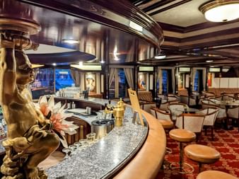 Luksuriøs bar ombord på MS Prinzessin Katharina med buet marmordisk, gyldne dekorative søjler, læder barstole og elegant siddeområde.