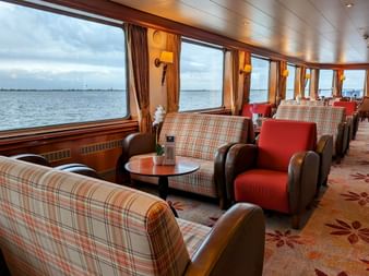 Interiør af MS Rigoletto salon med ternede og røde lænestole langs store vinduer med udsigt til vand, varm belysning og gulvtæppe.