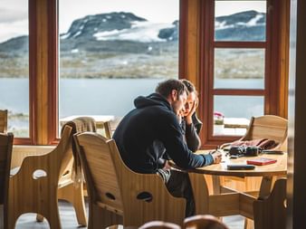 Person sidder ved træbord inde i DNT Finsehytta hytte og kigger ud gennem store vinduer mod sø og snedækkede bjerge i Hardangervidda, Norge.