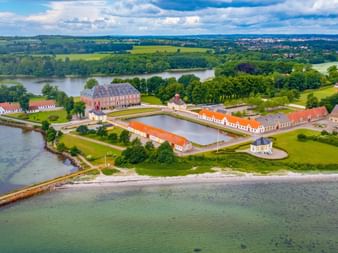 Flygbild av slottskomplexet Valdemars Slot på Tåsinge, Fyn, med rödtakade byggnader, gröna gräsmattor och läge vid kusten.