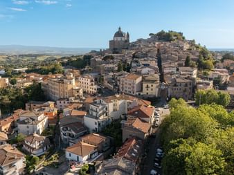 Aerial view of an Italian hilltop town with terracotta roofs and a prominent church dome. The town is surrounded by countryside on the Via Francigena.