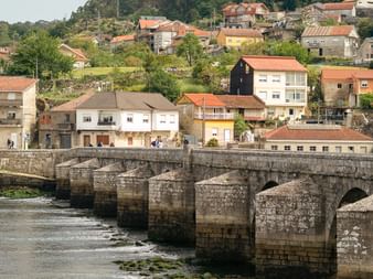 Historic stone bridge with multiple arches crossing a river in a Portuguese village. Houses with terracotta roofs line the hillside.