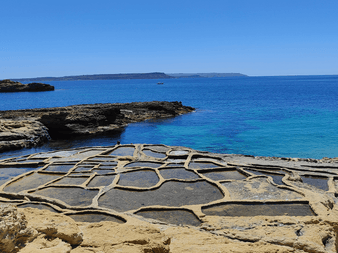 Traditionelle saltpander udskåret i kystklipperne på Gozo, Malta, med turkisblåt Middelhav og fjerne klipper under klar blå himmel.