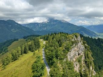 Winding mountain path along a green ridge with rocky outcrops in the Austrian Alps. Forested slopes and mountain peaks under cloudy skies.
