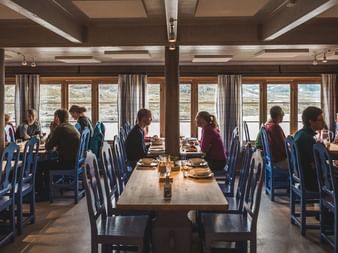 Interiør av spisestuen på DNT-hytta Geiterygghytta med vandrere ved trebord. Store vinduer viser fjelllandskap utenfor.