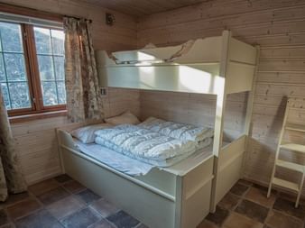 Hvid køjeseng med stige i et træhytteværelse ved Filefjellstuene. Naturligt lys kommer ind gennem et vindue med mønstrede gardiner.