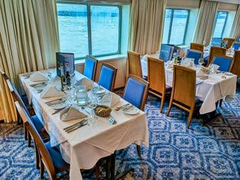 Elegant restaurant spisestue på MS SE-MANON med hvidt dækkede borde, blå polstrede stole og store vinduer med udsigt over vand.