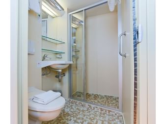 Kompakt badeværelse på MS SE-MANON med glasbruser, hvid vask, spejl, glashylder og mosaikgulv.