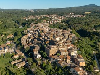 Aerial view of a historic Italian hilltop town with stone buildings and a church tower, surrounded by green valleys and forested hills.