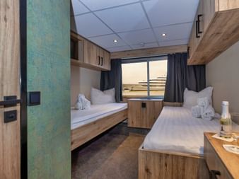 Moderne øvre dæk kahyt på MS PRINCESS med to enkeltsenge, træmøbler, stort vindue og blå gardiner. Hvide håndklæder på sengene.