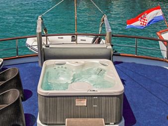 Firkantet spabad på blåt dæk på MS Azimut med kurvstole, metalgelænder og kroatisk flag over turkis vand.