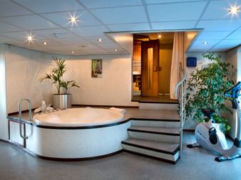 Moderne wellnessområde på MS SE-MANON med rundt spabad, træsauna, motionscykel og grønne planter under indbygget belysning.