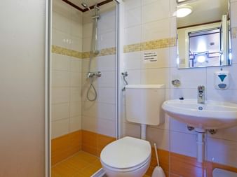 Lille skibsbadeværelse med glasbruser, hvidt toilet, vægmonteret vask og orange gulvfliser.