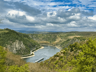 Luftfoto af Rhinen, der slynger sig gennem en grøn dal med stejle skråninger. Et fragtskib sejler på floden under en skyet himmel.
