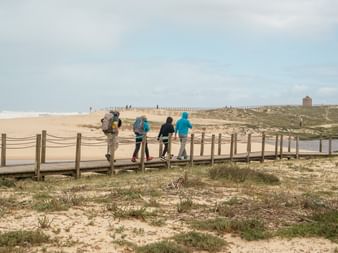 Four hikers with backpacks crossing a wooden footbridge over a stream on the Portuguese Camino, with sandy dunes and a windmill visible in the distance.