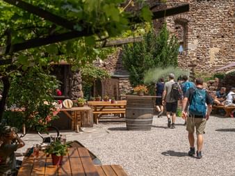 Gruppe vandrere med rygsække går gennem en rustik stengård med træborde, vintønder og traditionel italiensk arkitektur.