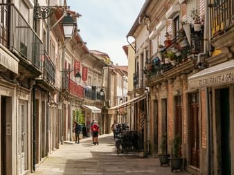 Narrow pedestrian street with traditional buildings and balconies. Two pilgrims walk along the stone-paved street lined with shops and restaurants.