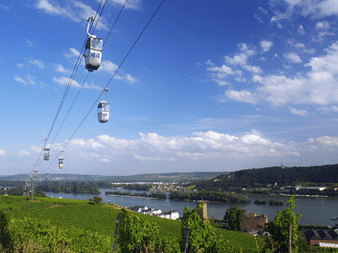 Svævebanegondoler over vingårde med udsigt over Rhinen nær Rüdesheim. Grønne vinrækker i forgrunden, flod og bakker i baggrunden.