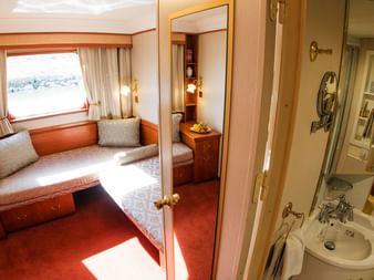 Flodkrydstogtskahyt på Donau med to senge, vindue med udsigt, træmøbler, rødt gulvtæppe og kompakt badeværelse med vask og bruser.