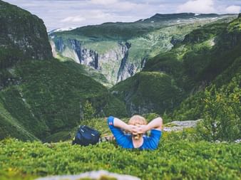 Turgåer i blå jakke hviler på gress med hendene bak hodet og ser utover grønn fjelldalføre i Aurlandsdalen, Norge.