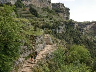To vandrere går på gamle stentrapper hugget ind i en bjergside ved Amalfikysten, omgivet af frodig grøn vegetation og kalkstensklipper.
